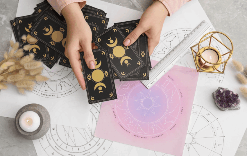 俯瞰占卜師的雙手正在整理帶有月亮週期圖案的黑色塔羅牌，桌面擺放著星座圖表、白色蠟燭、水晶簇以及乾燥植物裝飾