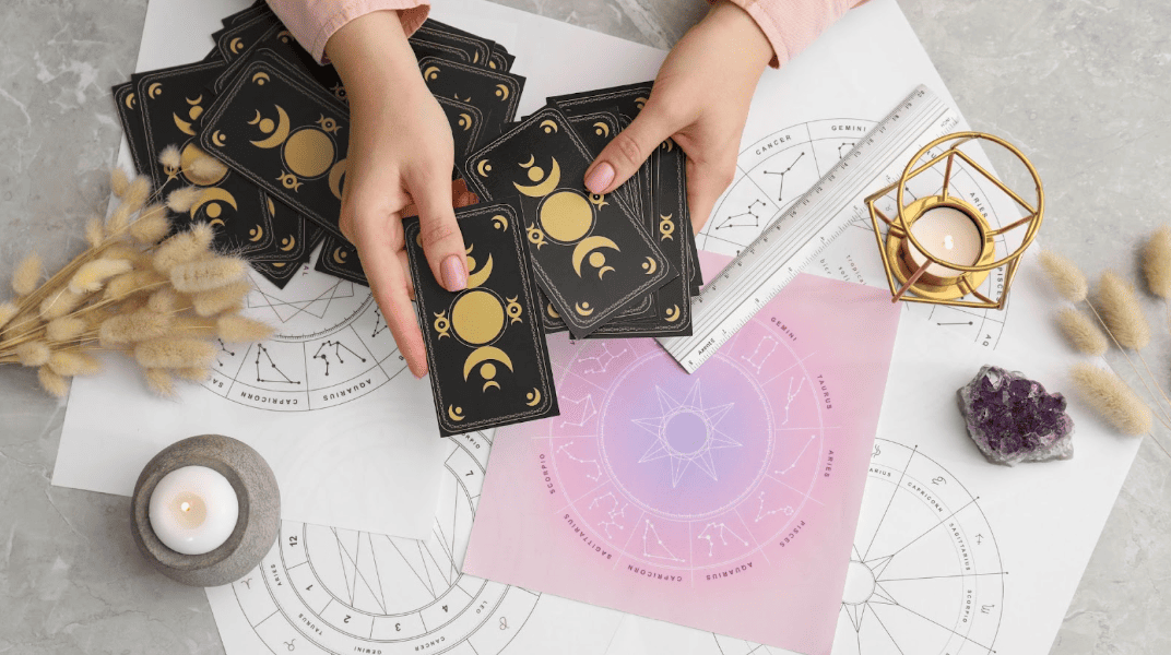俯瞰占卜師的雙手正在整理帶有月亮週期圖案的黑色塔羅牌，桌面擺放著星座圖表、白色蠟燭、水晶簇以及乾燥植物裝飾