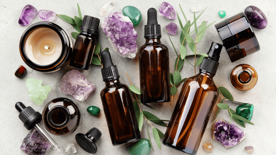灰色石紋背景上散布著多瓶深色玻璃精油滴瓶、紫水晶與綠色礦石，並點飾著尤加利葉與點燃的蠟燭，營造芳香療法氛圍