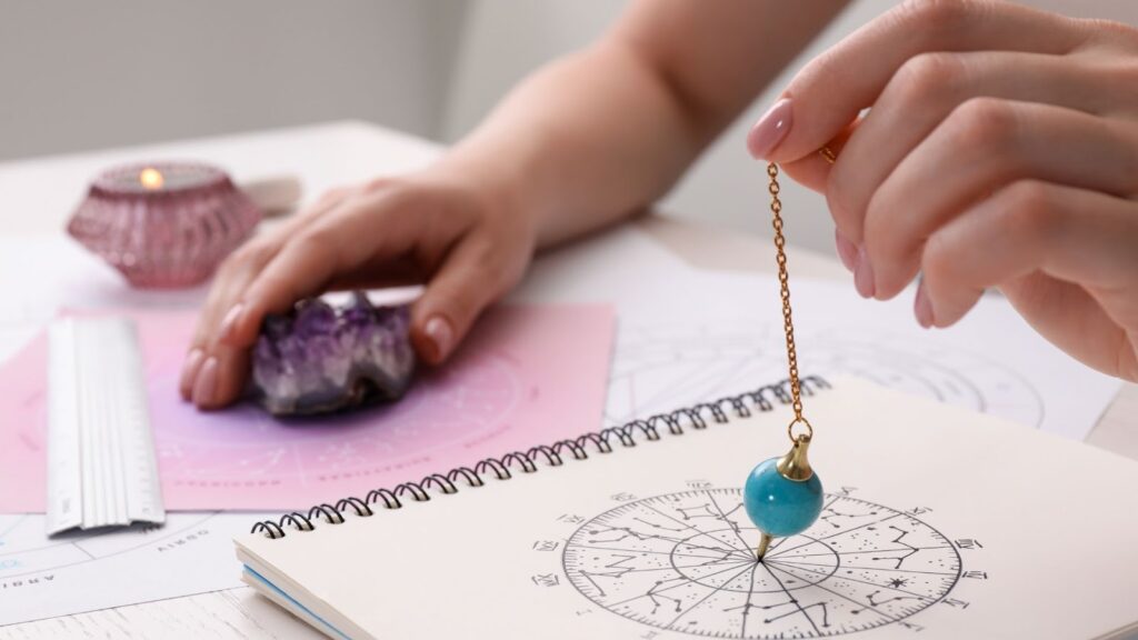 占卜師手持藍色礦石靈擺，在筆記本上的星盤圖案上方進行測量，背景可見紫水晶、星座盤以及點燃的粉色蠟燭。