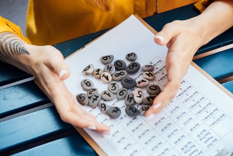  一名女子張開雙手置於桌面上，桌上散落著寫有數字的小圓石與一張生命靈數分析表格，呈現出神祕學占卜與命理諮詢的場景。 