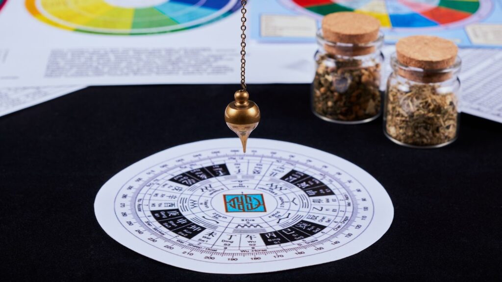 占卜桌面上的金色靈擺正懸掛在圓形易經八卦方位圖上方，背景有兩罐乾燥草藥、色彩分析圖表及黑色布面。