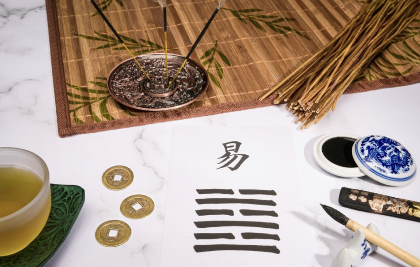 一張白色宣紙上寫著「易」字與八卦卦象，周圍擺放著古代銅錢、毛筆、墨條、茶杯與點燃的線香，營造出濃厚的傳統易經占卜氛圍。