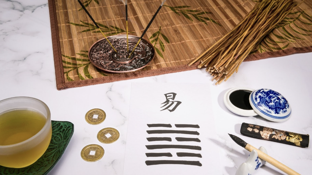 一張白色宣紙上寫著「易」字與八卦卦象，周圍擺放著古代銅錢、毛筆、墨條、茶杯與點燃的線香，營造出濃厚的傳統易經占卜氛圍。