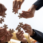 由下往上的仰拍視角，多位穿著西裝的人員在藍天雲朵下，共同拿著棕色拼圖塊並嘗試將其組合在一起，象徵團隊合作