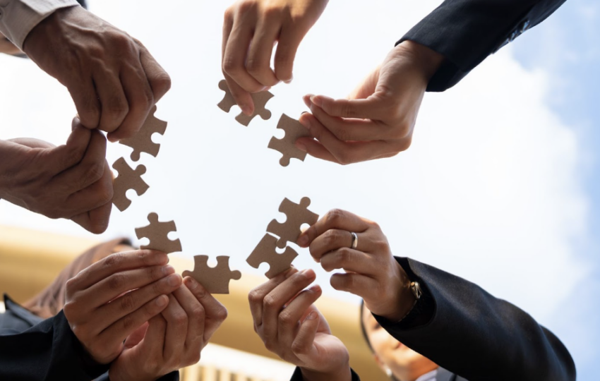 由下往上的仰拍視角，多位穿著西裝的人員在藍天雲朵下，共同拿著棕色拼圖塊並嘗試將其組合在一起，象徵團隊合作