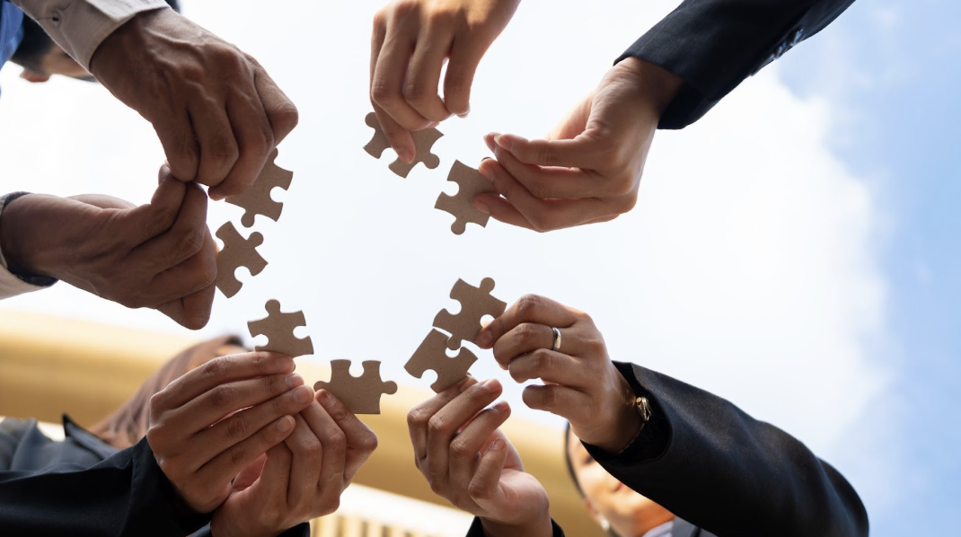由下往上的仰拍視角，多位穿著西裝的人員在藍天雲朵下，共同拿著棕色拼圖塊並嘗試將其組合在一起，象徵團隊合作